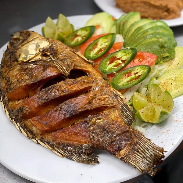 Crispy fried whole fish with sliced avocado, jalapeños, tomatoes, and lime wedges on a white plate.