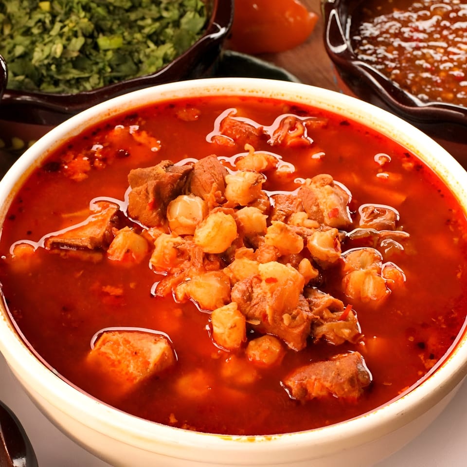 Close-up of a bowl of red pozole with hominy and pork, with cilantro and salsa in the background.