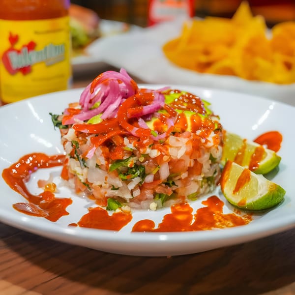 Mexican ceviche with red onions, avocado, hot sauce, and lime wedges on a white plate.