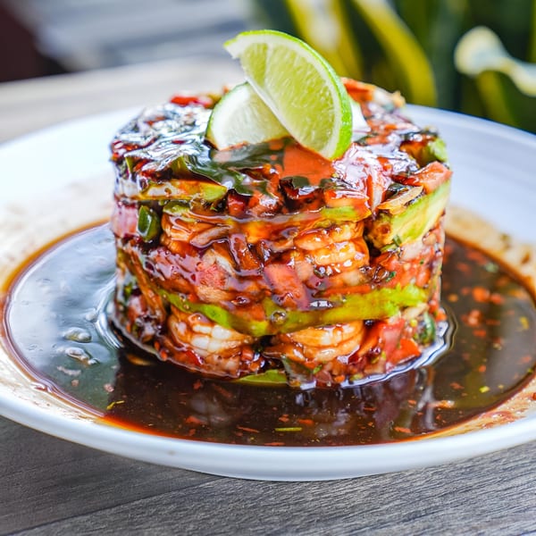 Shrimp ceviche stack with avocado, tomatoes, and lime wedges, drizzled with dark sauce on a white plate.