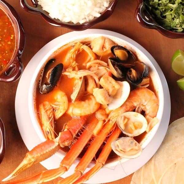 Overhead view of a vibrant seafood soup with crab legs, shrimp, mussels, and clams, served with tortillas and lime.