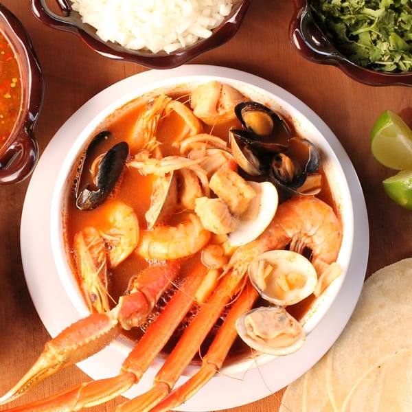A large bowl of Mexican seafood soup with crab legs, shrimp, mussels, and clams, with sides.
