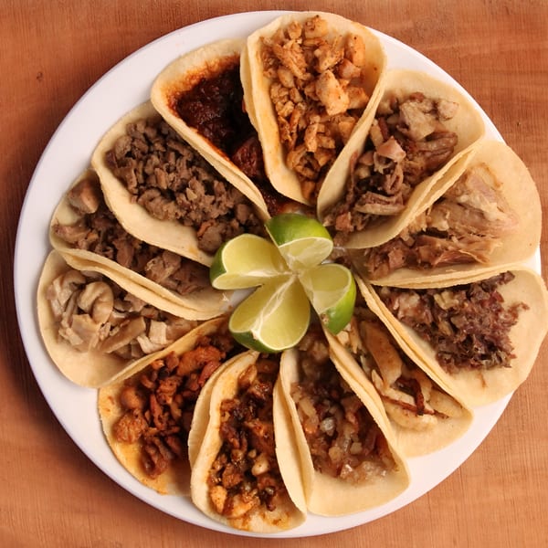 A white plate holds a dozen Taqueria Los Portales tacos with various fillings and lime wedges.