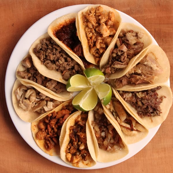 A white plate filled with various Mexican street tacos, garnished with lime wedges, on a wooden surface.