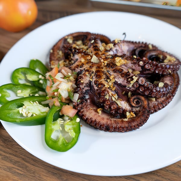Grilled octopus with garlic, pico de gallo, and sliced jalapeños on a white plate.